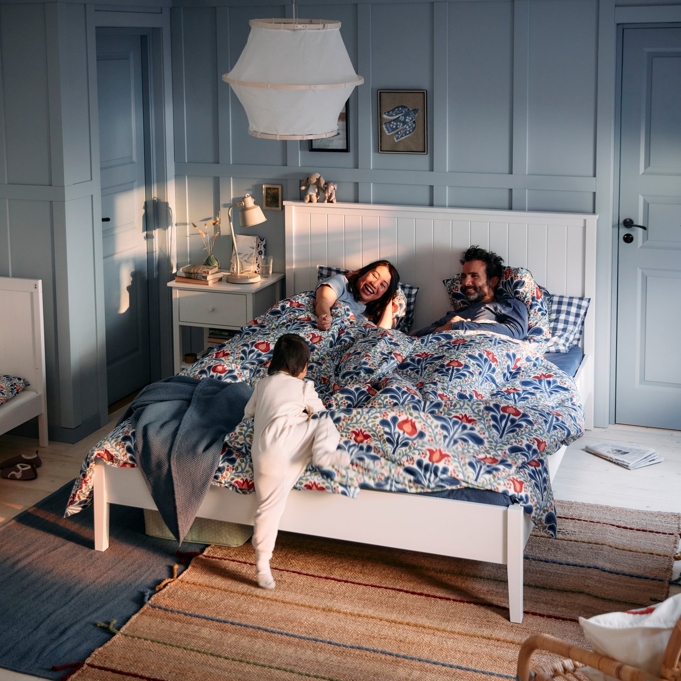 Chambre avec housse de couette SÄTERVIDE au motif fleuri sur un lit blanc où deux personnes et un enfant jouent.