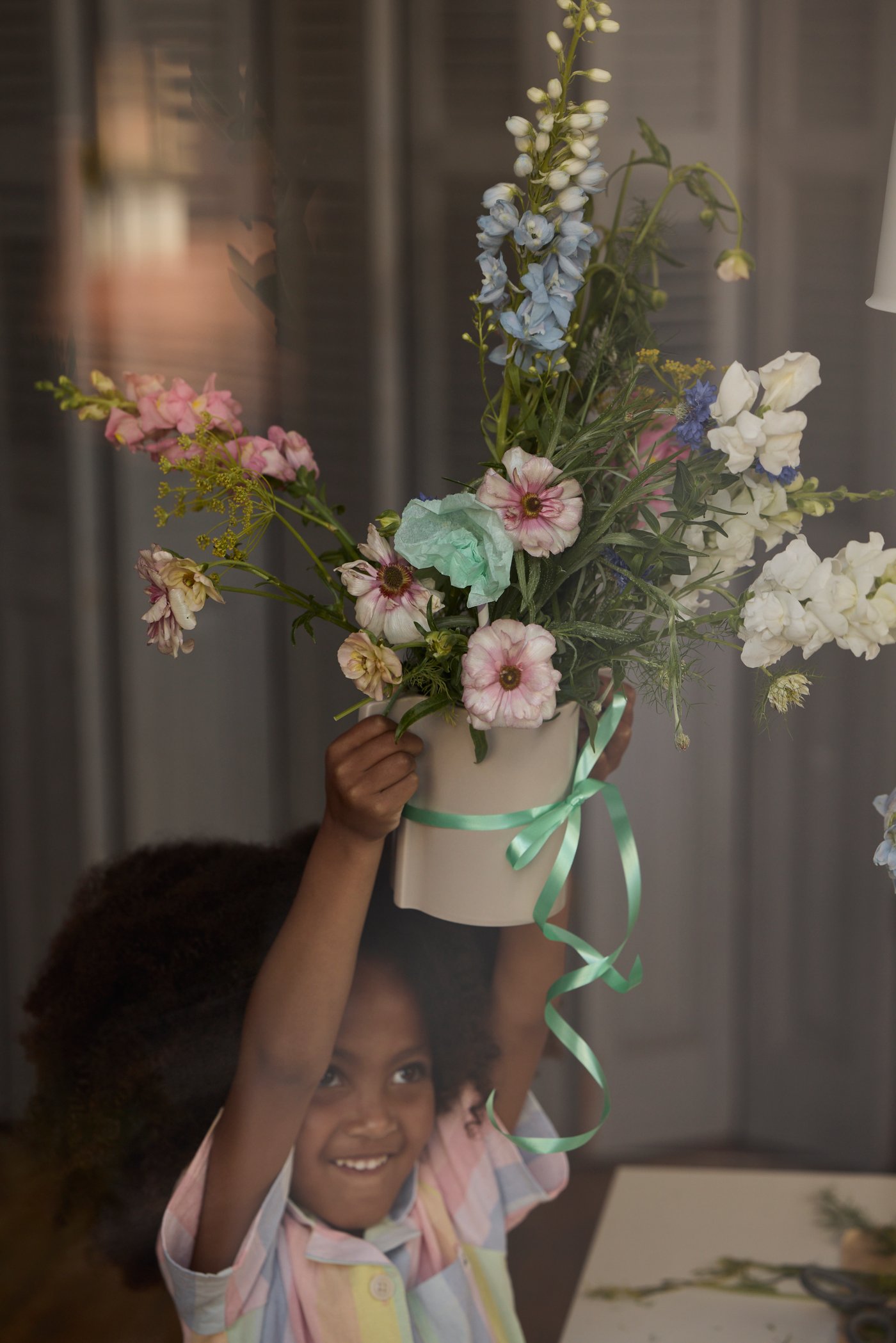 A smiling child holds up a beautiful bouquet of fresh spring flowers, including pink, blue, and white blossoms, in a pot decorated with a green ribbon.