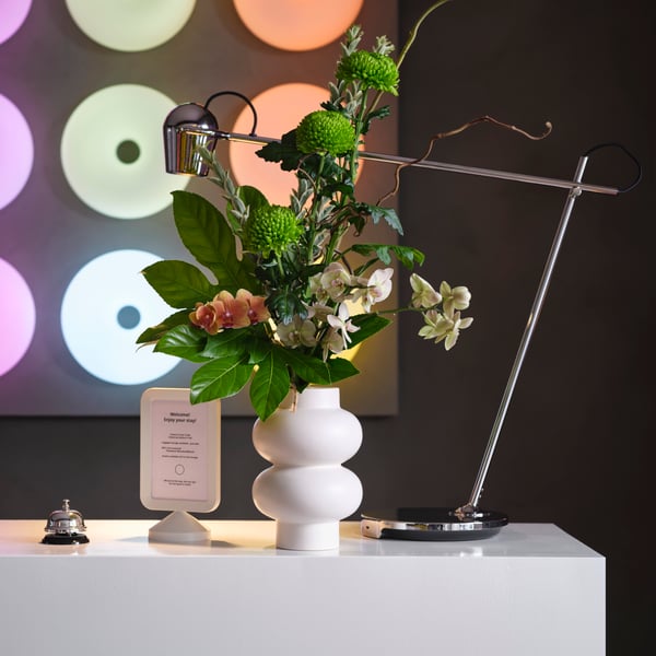 A curvy, beige stoneware PÄRLHÄGG vase holds fresh flowers on a white hotel reception desk, by a lamp, a bell and signage.