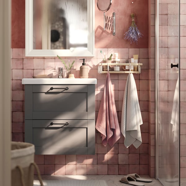 Un meuble lavabo avec tiroirs HAVBÄCK gris et un meuble à miroir au-dessus, dans une salle de bain aux murs rose tendre et au sol gris.