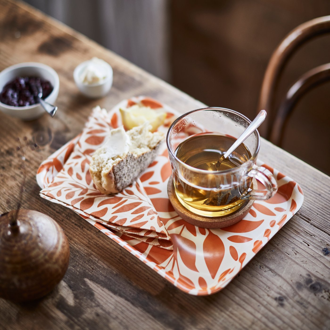 A HÖSTAGILLE tray with an orange pattern displays a slice of buttered bread, a glass of tea, and a HÖSTAGILLE paper napkin.