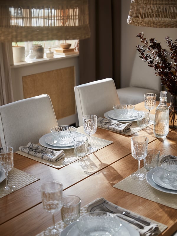 On a table are place settings with placemats, plates, glass bowls, glasses, wine glasses, cutlery and linen napkins.