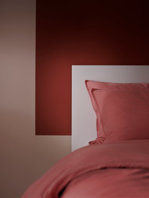 Vue partielle d’un lit habillé de linge de lit ÄNGSLILJA brun-rouge, contre un mur peint partiellement dans un ton brun-rouge assorti.
