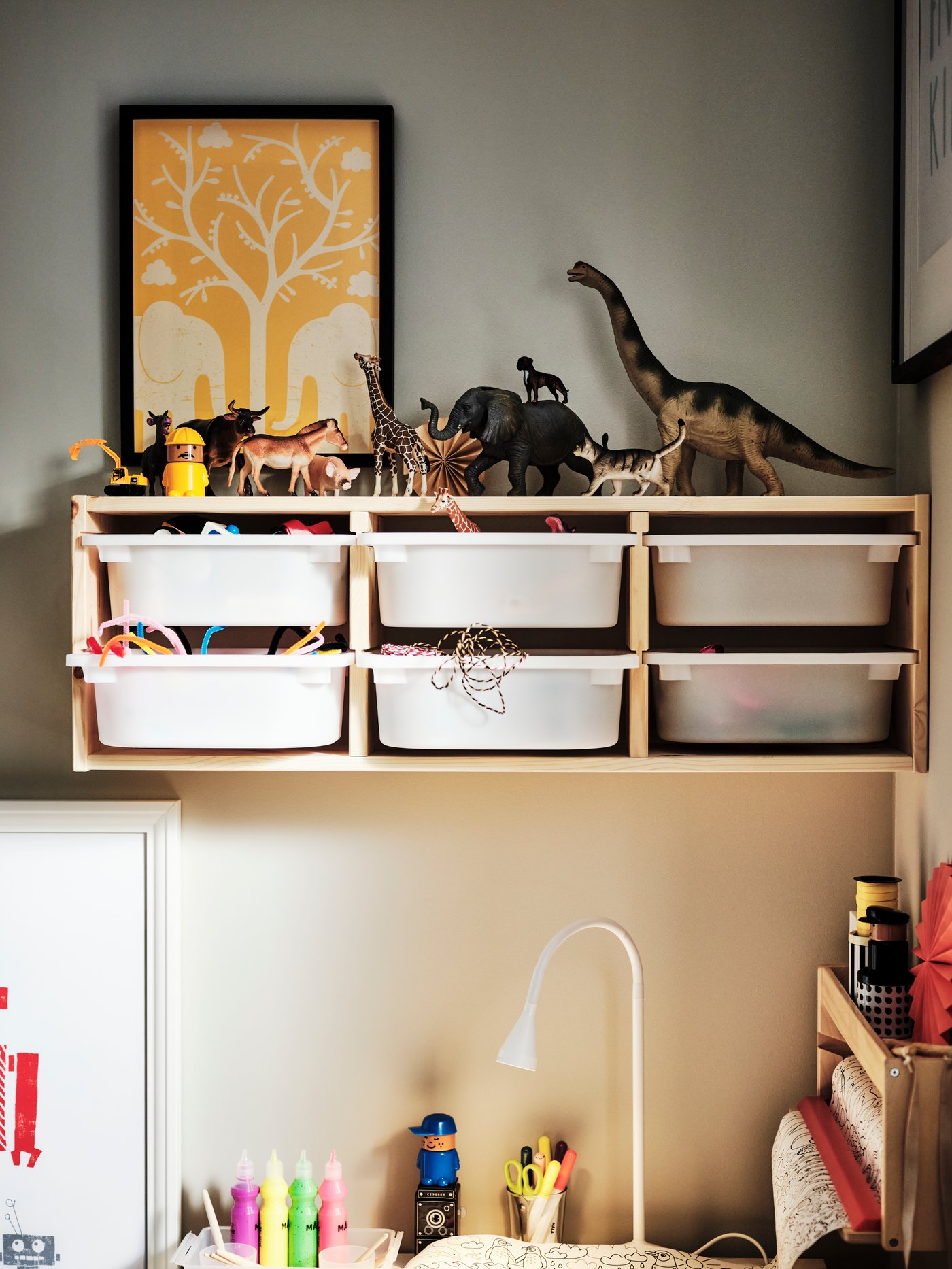 Une chambre d’enfant ou coin jeu avec une étagère remplie de bacs blancs, des jouets dinosaures posés dessus, une œuvre orange au mur et un bureau en dessous avec des feutres et une lampe.