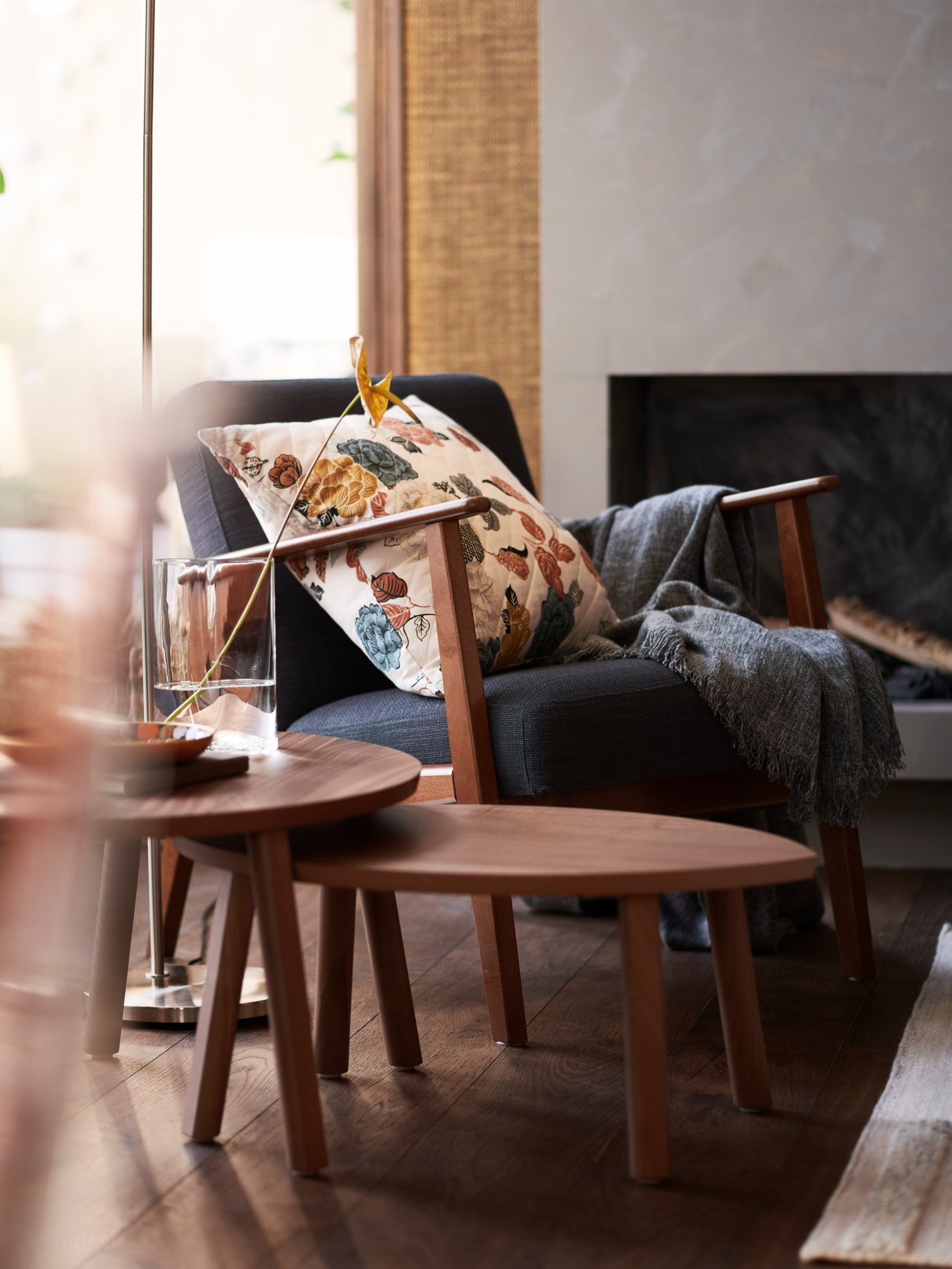 An EKENÄSET armchair with a dark grey cover and cream cushion by a window in a living room, with coffee tables beside it.