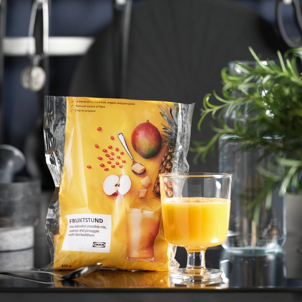 A kitchen worktop with a bag of FRUKTSTUND mango and pineapple smoothie next to a glass of smoothie and a tablespoon.
