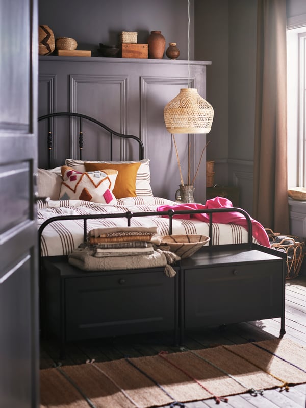 Two black storage benches and a bed with a black bed frame made with striped bed linen and a BRUNSTARR multicolor cushion.