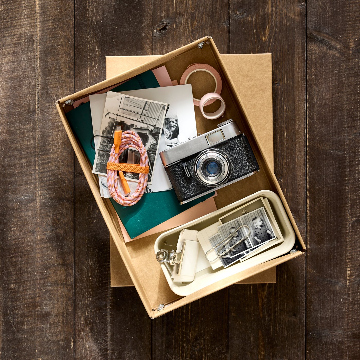Una caja de almacenamiento de papel GÄRDESGÅRD, sin la tapa, está colocada sobre un piso de madera y contiene accesorios de fotografía.