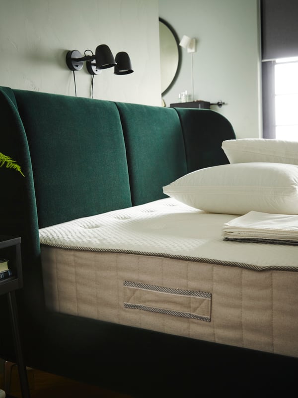 A green TUFJORD bed with an uncovered VATNESTRÖM mattress and pillows on it. Two SKURUP wall lights are behind the bed.