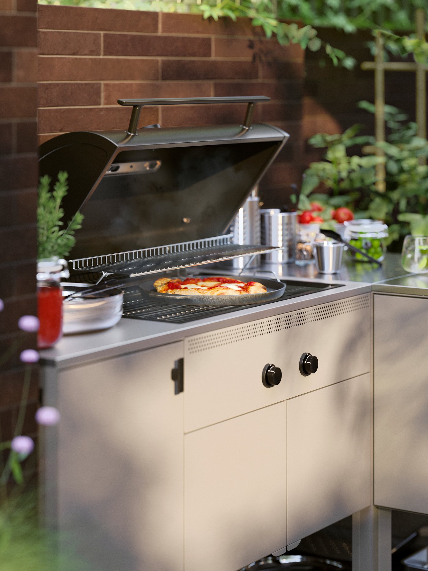 A patio in evening light and a pizza baking on a BÅTSKÄR outdoor kitchen with gas barbecue next to herbs and tomatoes.