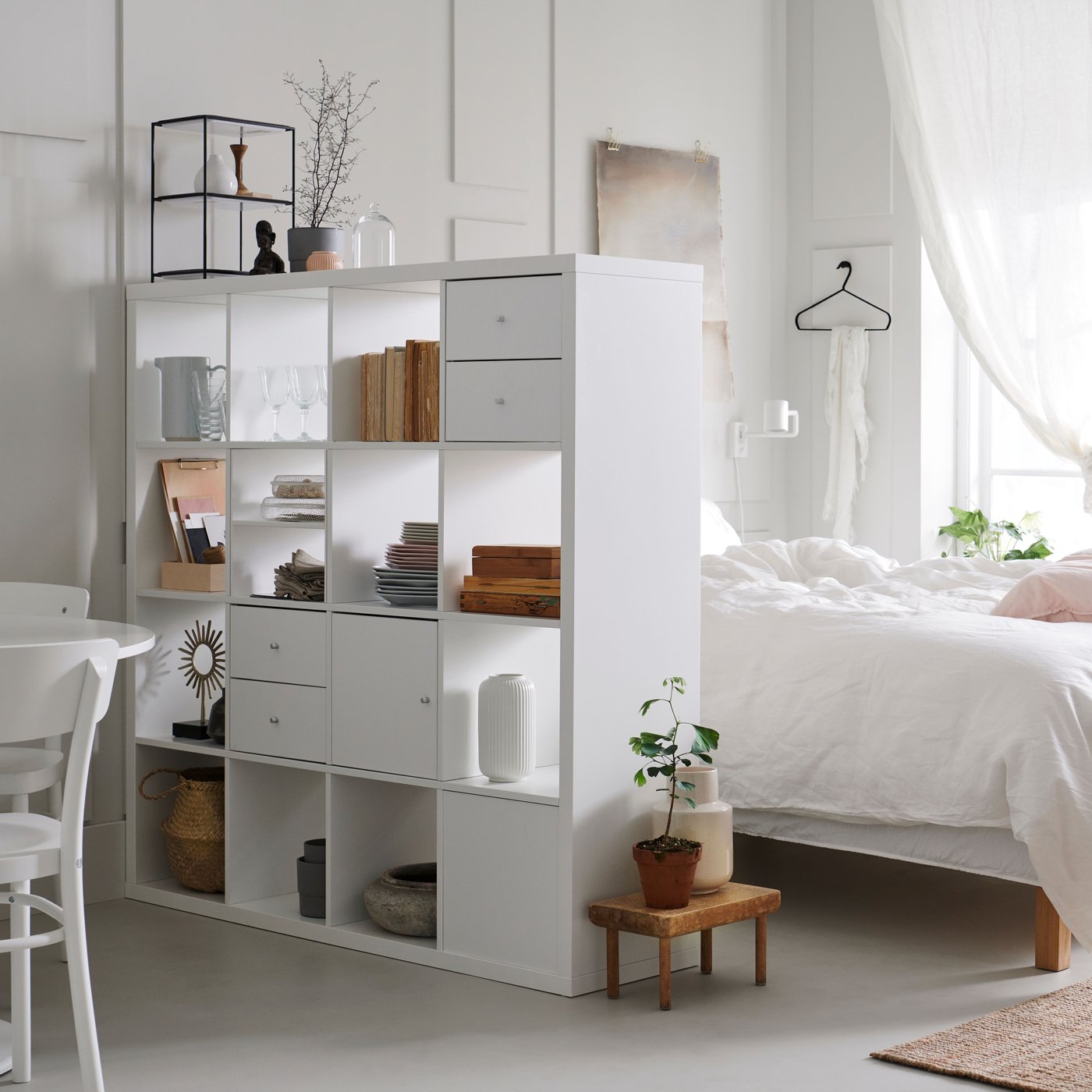 A bedroom featuring a white KALLAX shelving unit filled with decor and storage boxes next to a double bed with white linen.
