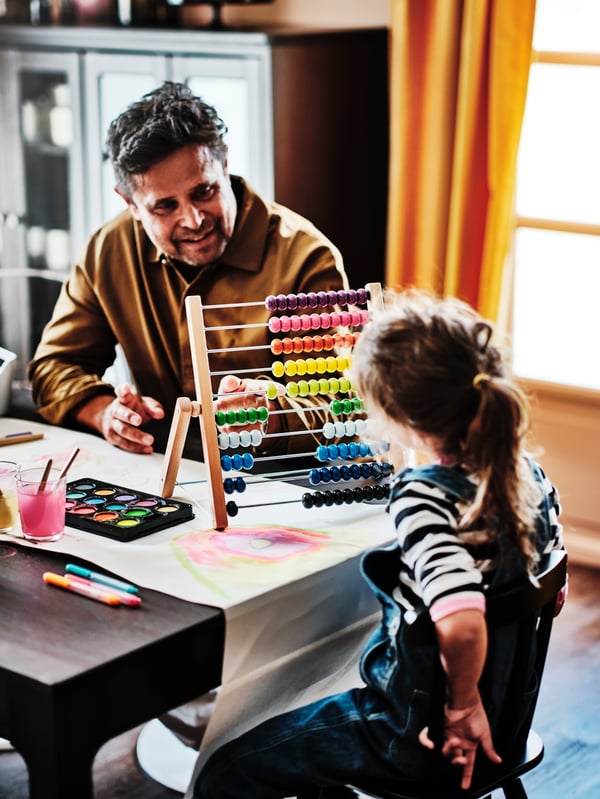 An adult and child sitting at a table with a multicolour UNDERHÅLLA abacus between them on the table.