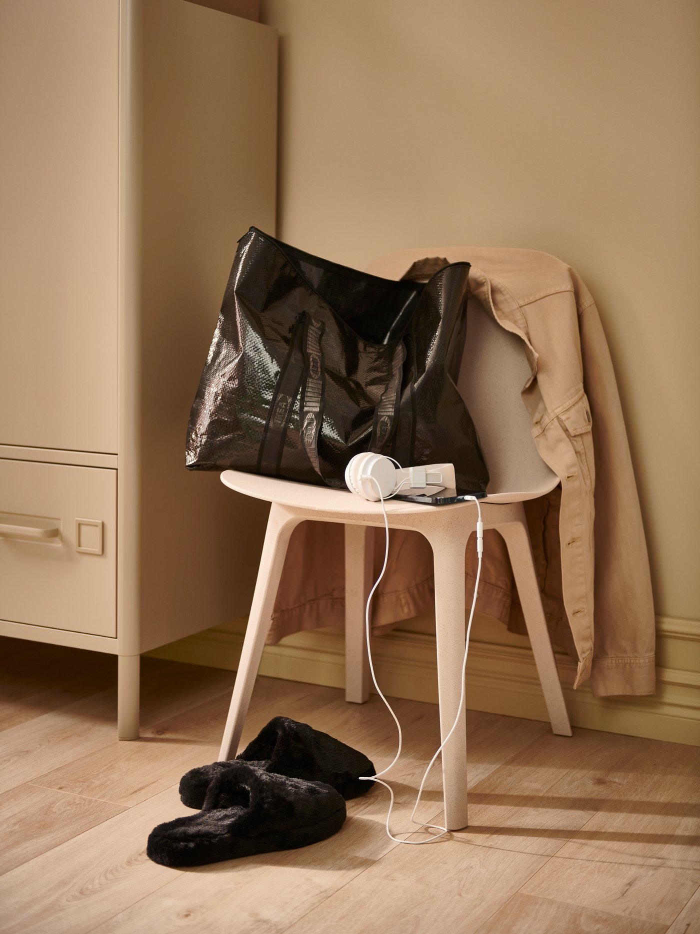 A black OBEGRÄNSAD bag and slippers with white headphones and beige jacket on a chair next to a beige cabinet.