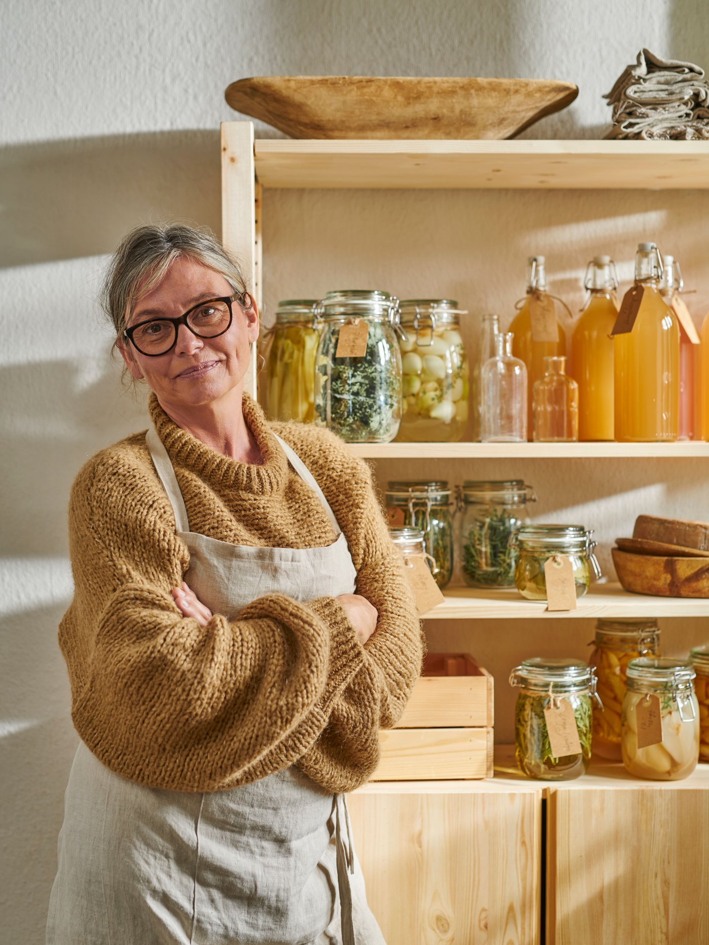 A person, wearing glasses, a mustard-coloured sweater, and an apron, is standing next to a shelf with bottles and jars.
