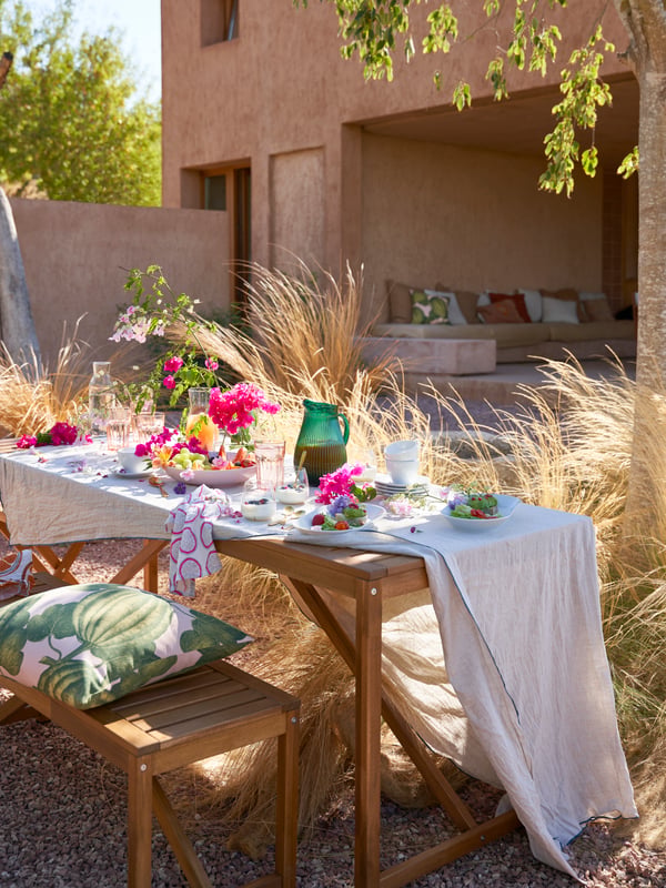 A light brown stained outdoor NÄMMARÖ bench and table laid with glasses, plates and flowers in a clear glass BERÄKNA vase.