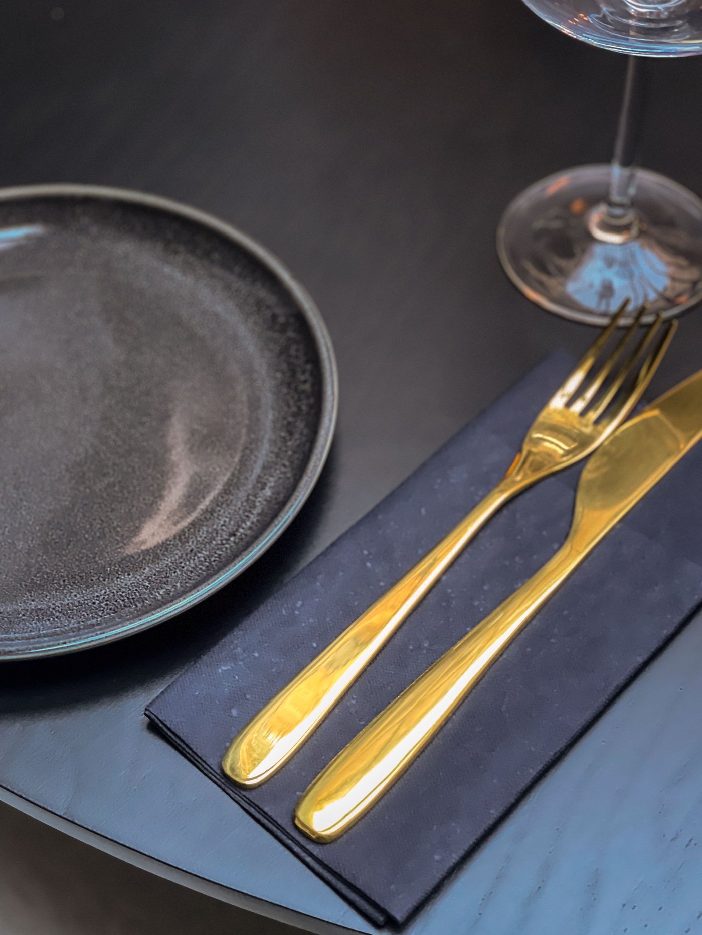 Donkere tafel met zwart bord, gouden bestek op servet en wijnglas. Sfeervol en elegant gedekt voor een verfijnde restaurantbeleving.