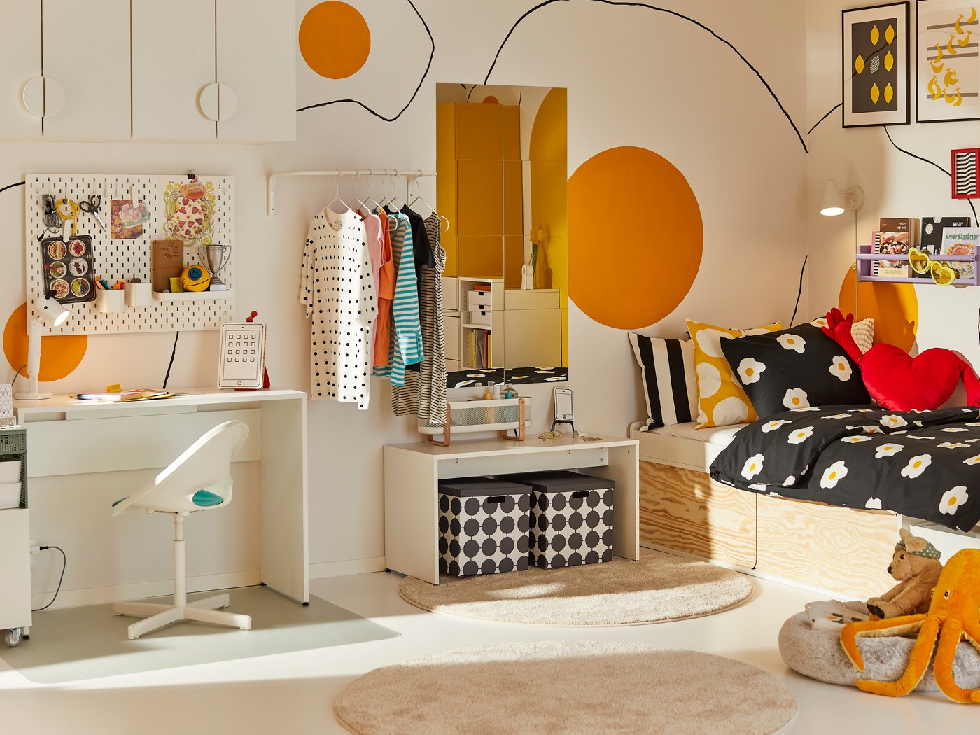 A bright, modern bedroom with a white desk, colorful clothes rack, and playful orange wall circles. The bed has a black and white daisy duvet with red heart pillows. Storage bins and a round rug complete the cozy space.