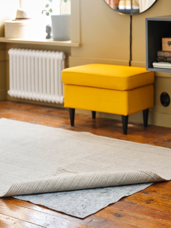 A corner of a TIPHEDE rug lying on a floor is pulled back to show STOPP FILT rug underlay with anti-slip underneath it.