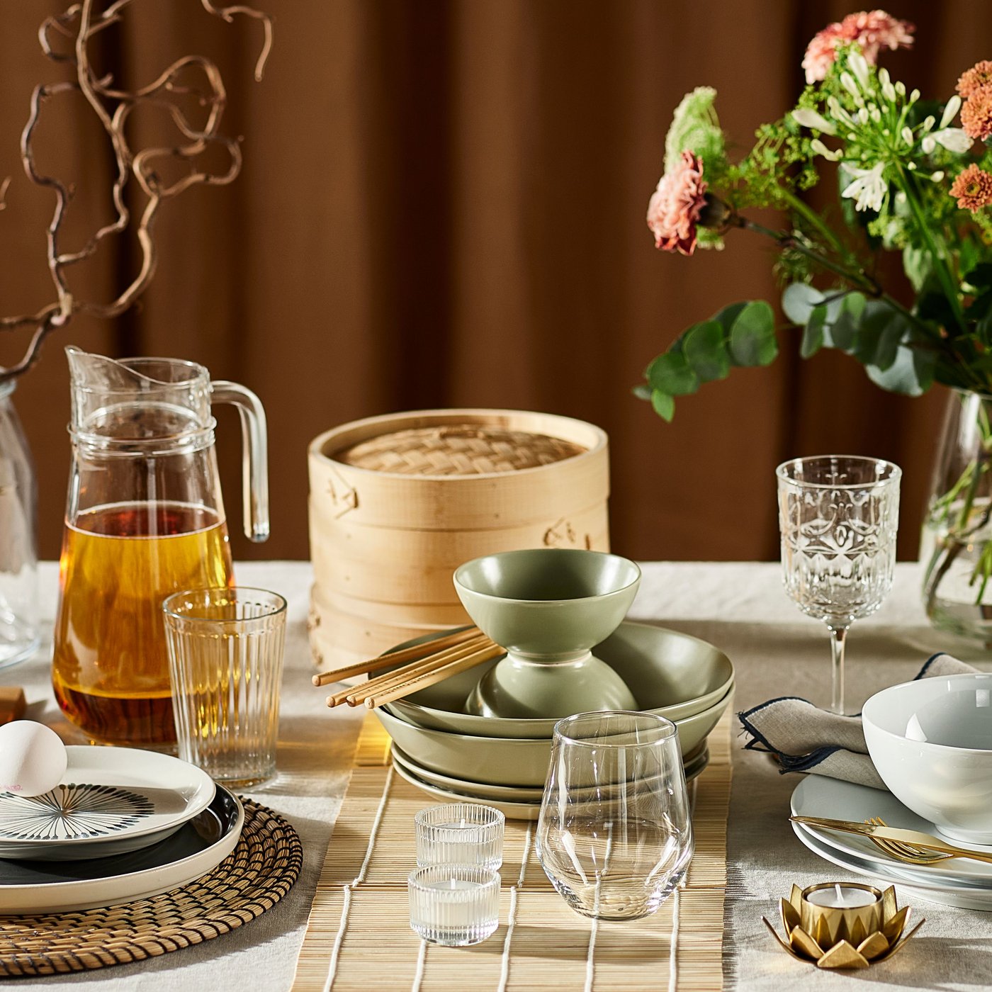 Part of a table with lots of items on it, such as flowers, glasses, matte green FÄRGKLAR plates, and a TILLBRINGARE jug.