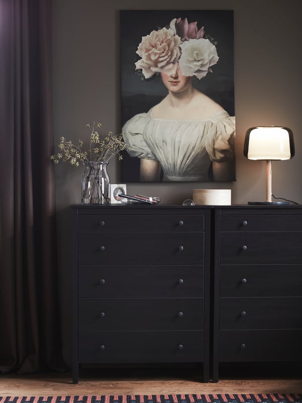 A KOPPANG chest of drawers in dark brown with an EVEDAL table lamp on it, in front of a wall-mounted painting.