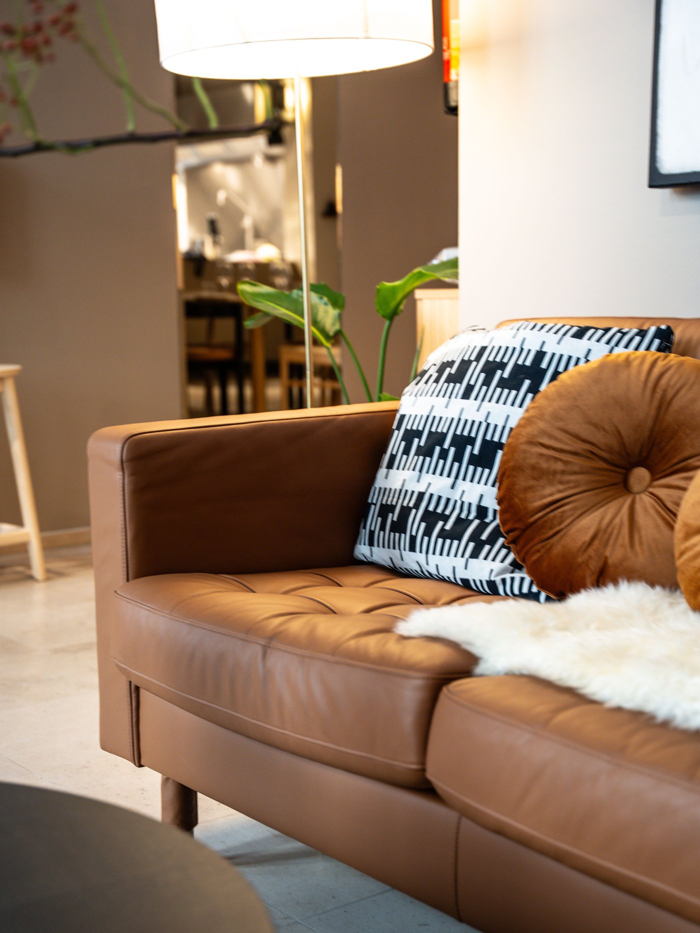 Bruine lederen zetel met kussens en plaid in warme zithoek. Op achtergrond plant, lamp en zicht op eetruimte met houten tafel en stoelen.