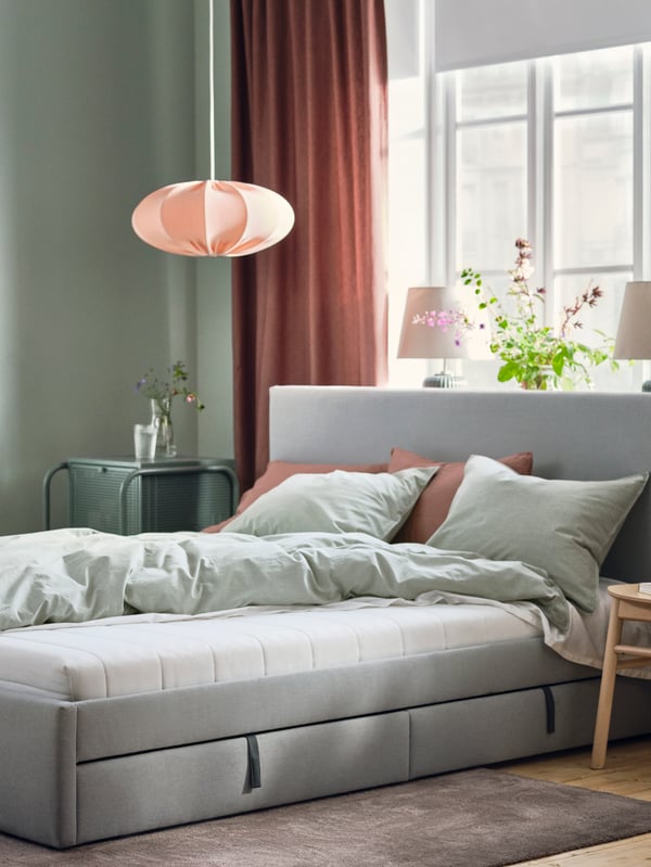 An upholstered bed in a light green bedroom in front of a window with dishevelled bed linen showing an ÅSVANG foam mattress.
