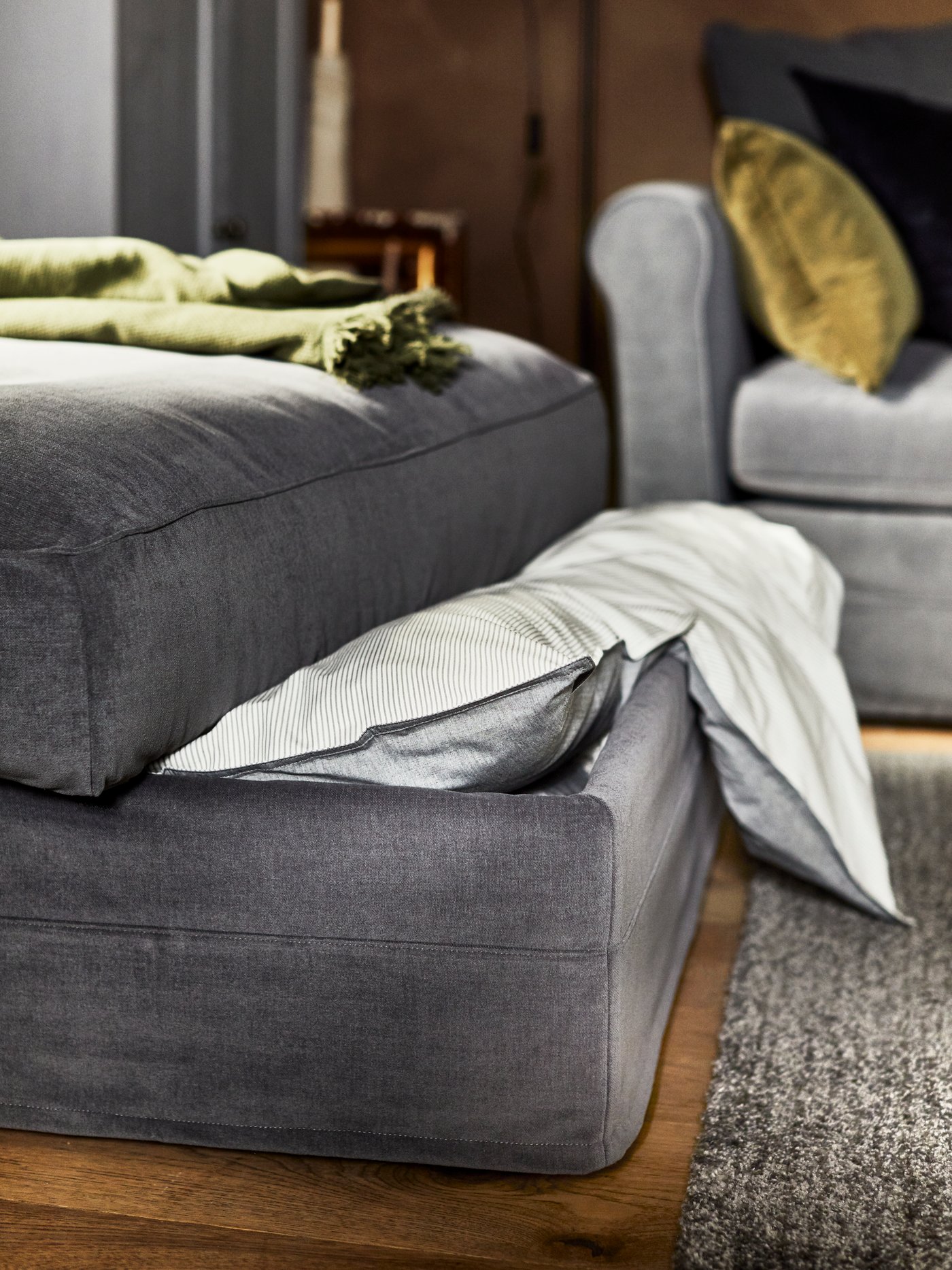 A grey GRÖNLID footstool with storage partly open with the edge of a duvet peeking out stands in a living room.
