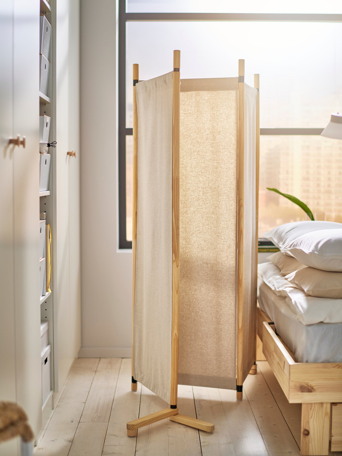 En GLAMBERGET rumsavdelare i obehandlat trä och beige textil står mellan säng och garderob. Ett stort fönster finns bakom sängen.
