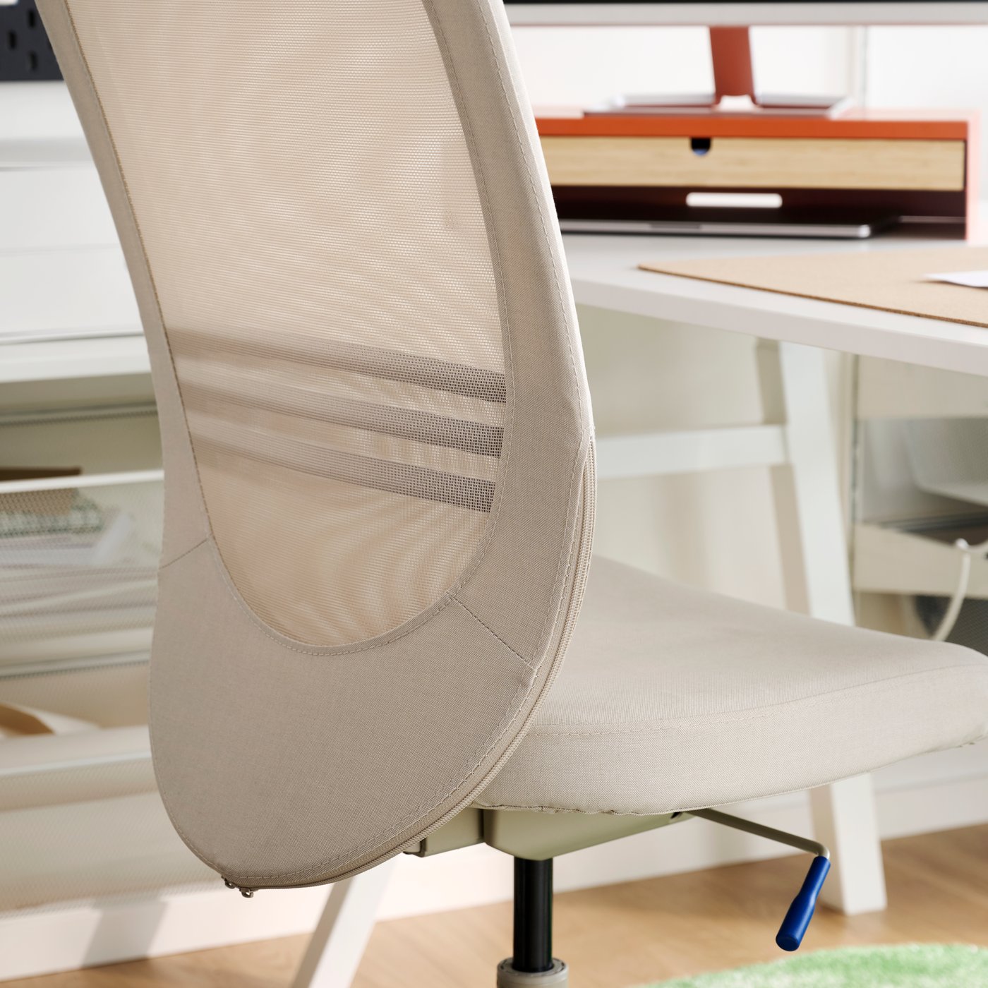 Acercamiento de una silla de trabajo FLINTAN beige con respaldo de malla junto a un escritorio en un espacio de oficina luminoso.