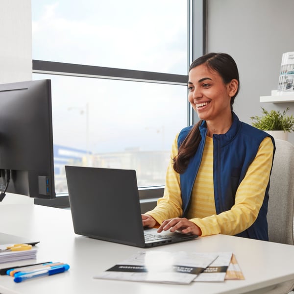 Una persona sorridente con un pullover giallo e un gilet blu lavora su un laptop nero appoggiato su una scrivania bianca accanto a una finestra.