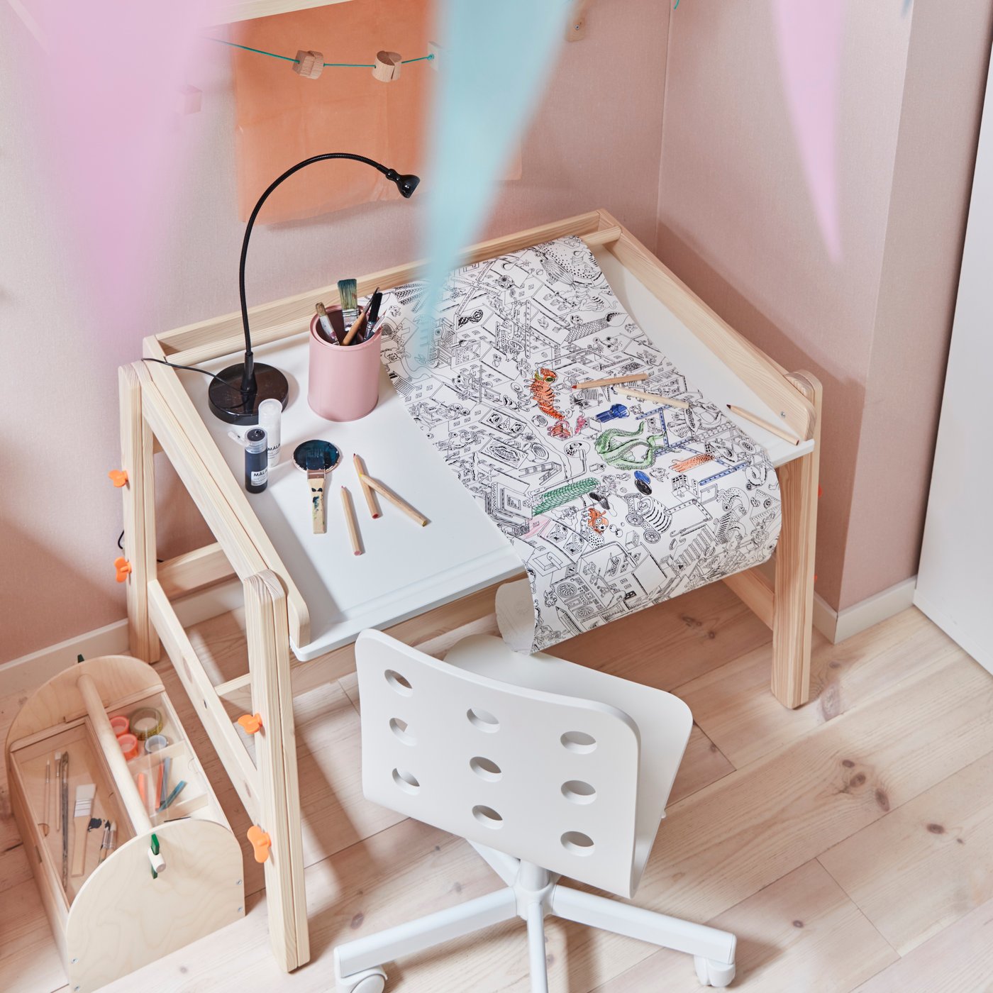 Un escritorio infantil FLISAT regulable y una silla infantil JULES ubicados en la esquina de un recinto con muros de color rosado claro. Sobre el escritorio se ven útiles para manualidades infantiles y una lámpara de trabajo. 