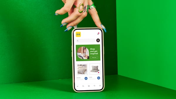 Ruka s noktima nalakiranim u zelenu boju drži pametni telefon na kom je prikazana IKEA aplikacija naspram zelene pozadine.