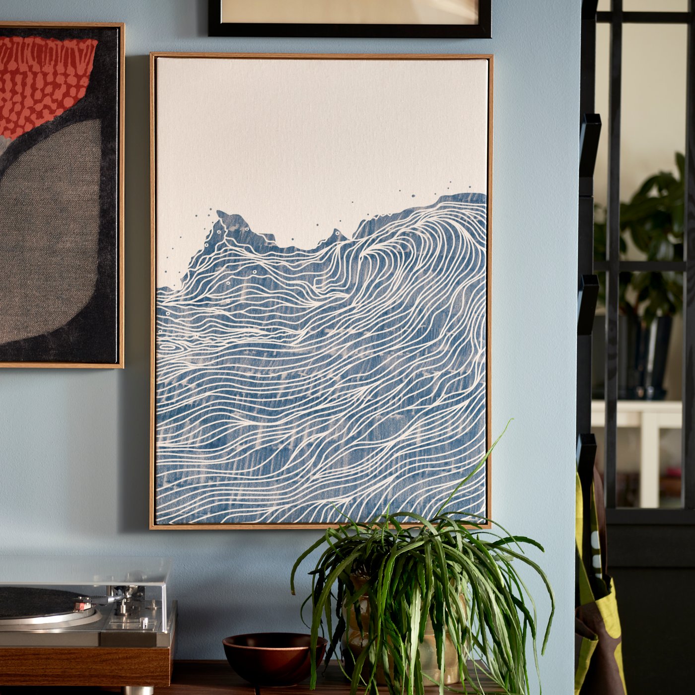 A light-blue wall section where framed BLODFLÄDER pictures hang side by side above a leafy potted plant and a gramophone.