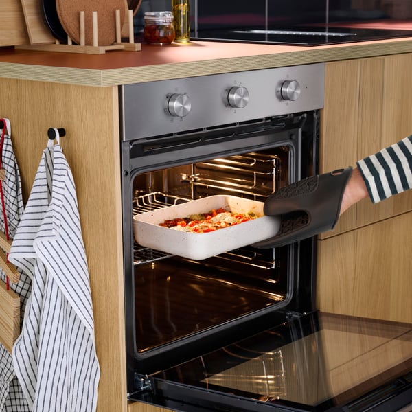 A person in a blue and white striped shirt is taking out a casserole from a FRILLESBO fan oven in a stainless steel colour.