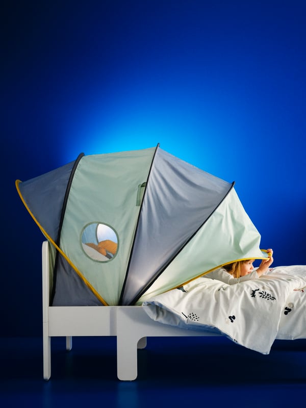 A blue/green ELDFLUGA bed tent on a SLÄKT bed against a blue wall. A child on the bed pulls the tent forward to close it.