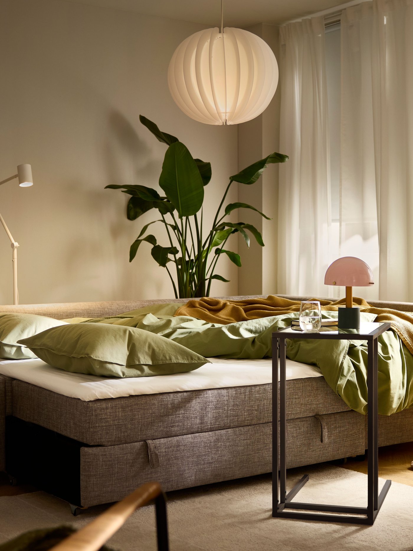 A NÖDMAST lamp on a VITTSJÖ laptop stand by a FRIHETEN sofa-bed in bed mode, bedding undone, in a softly lit living room.