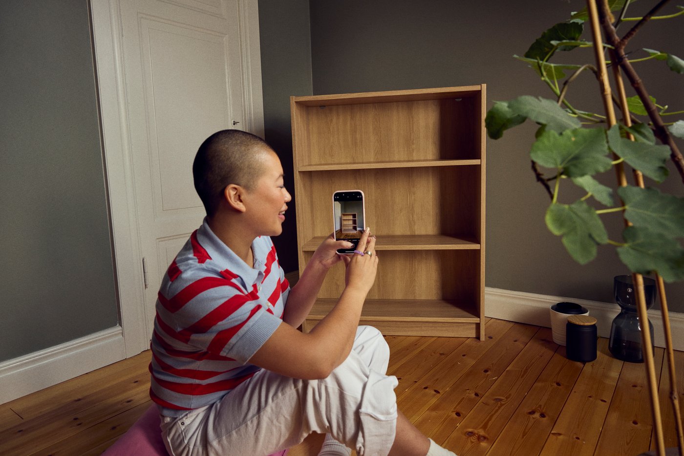 En person sitter på golvet i ett rum och fotograferar en öppen träbokhylla med sin mobiltelefon, för att lägga ut det på IKEA second-hand marketplace.
