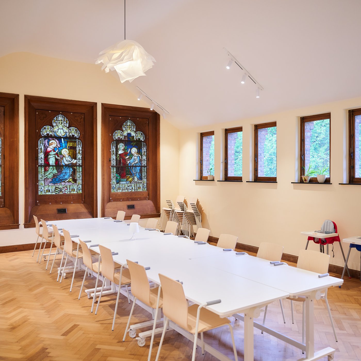 Salle de réunion lumineuse avec tables modulables et chaises, vitraux historiques, parquet et grandes fenêtres donnant sur la verdure.