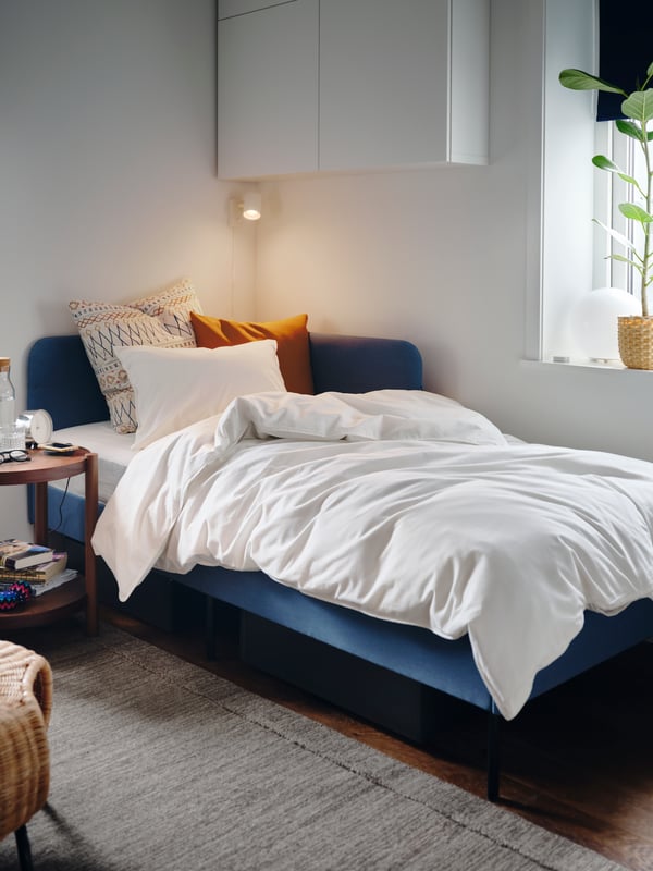 Una estructura de cama Knisa tapizada en azul medio con cabecero de esquina, funda de edredón, funda de almohada y cojines blancos.
