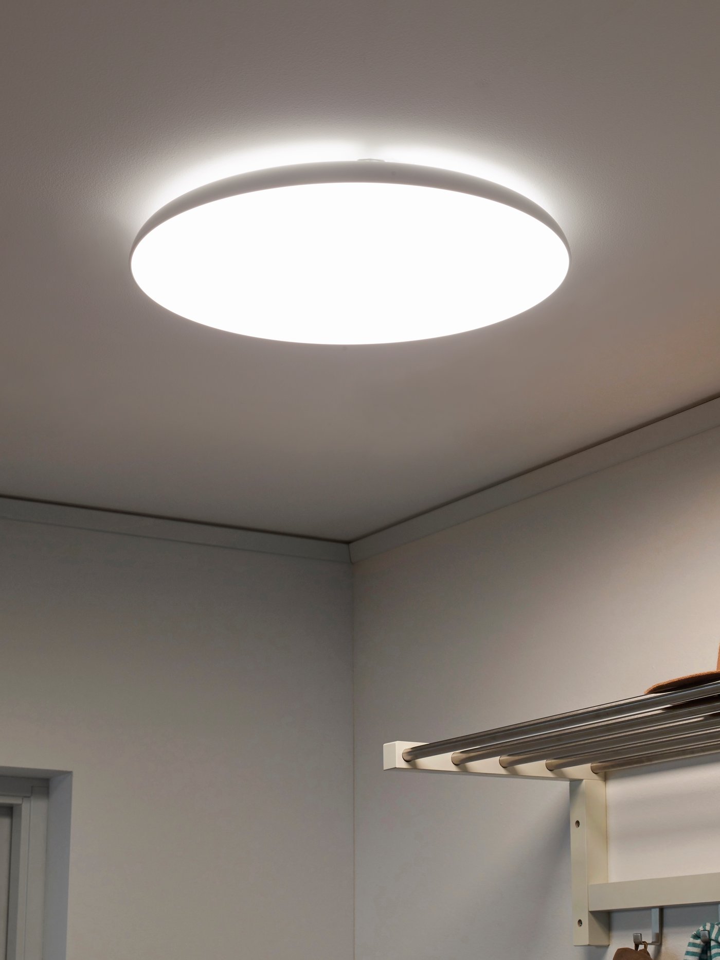 A round white NYMÅNE ceiling lamp that’s lit in the corner of a room near a door frame and with a shelf on the wall below.