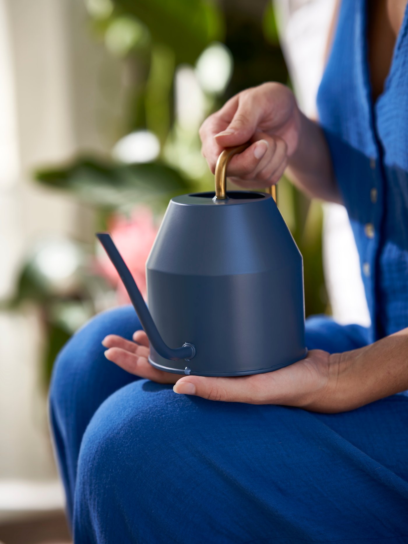 The hands of a person holding a dark-blue-and-gold-coloured VATTENKRASSE watering can, in front of leafy plants.