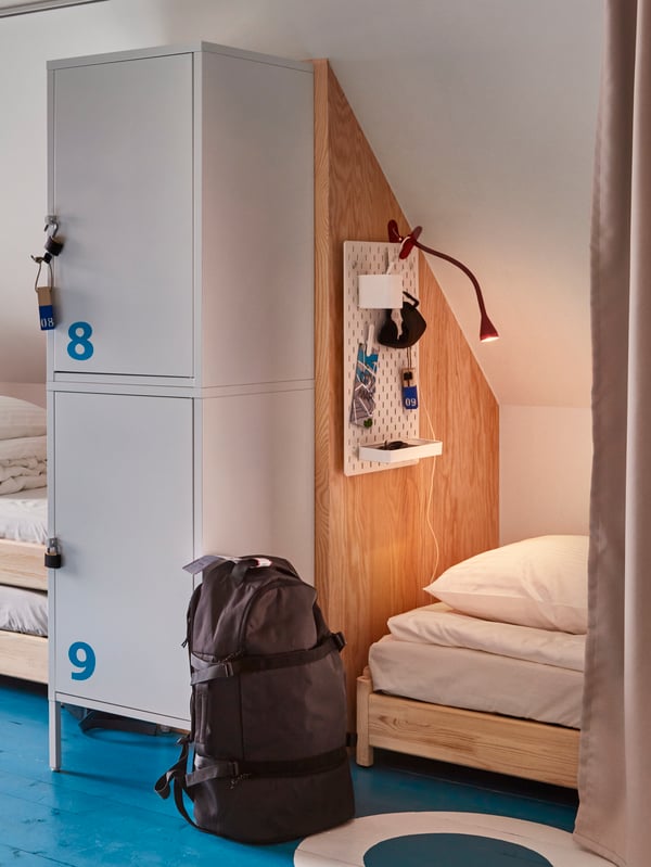 Delovi dva kreveta u hostelu, razdvojeni zidom i ormarima za odlaganje, ispred kojih je crni ranac.