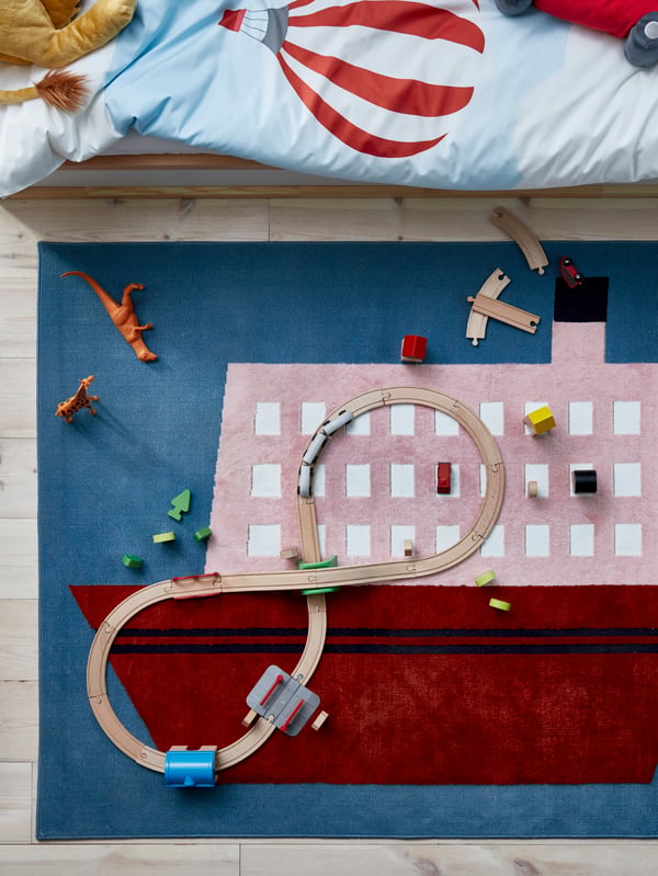 An UPPTÅG rug with a large boat motif lies near a bed. Toys including a wooden train set have been placed on the rug.