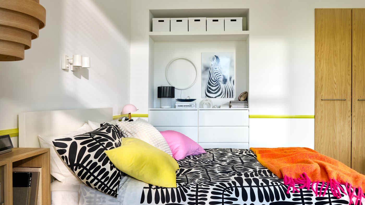 A bedroom with white walls, built-in storage in the back and a double bed with bright pink and black/white bedding.