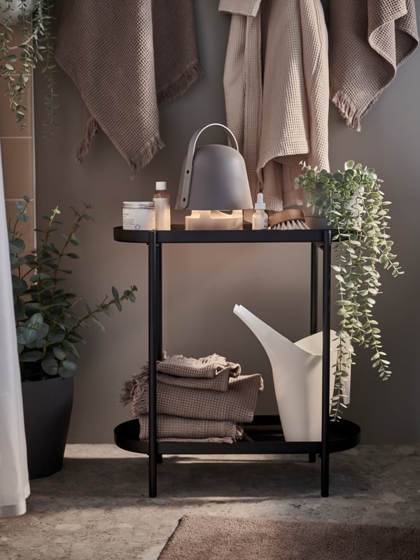 Un piédestal noir dans une salle de bains sur lequel est posé une lampe haut-parleur Bluetooth VAPPEBY d’extérieur/grise et divers autres objets.