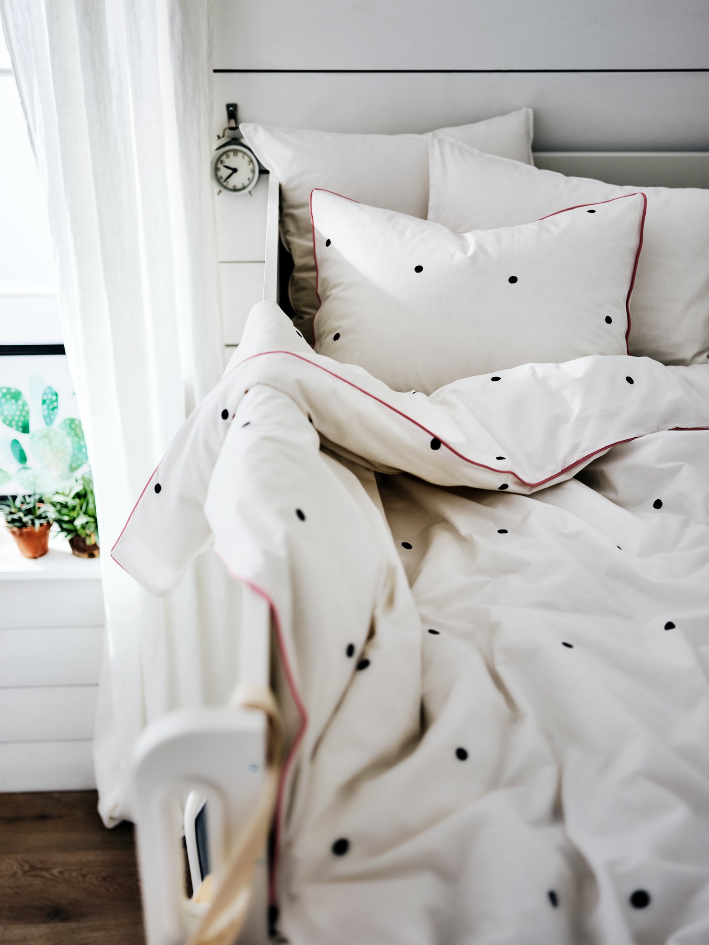 La partie supérieure d’un lit mezzanine devant une fenêtre avec des draps de lit blancs à pois noirs et un bord rouge.
