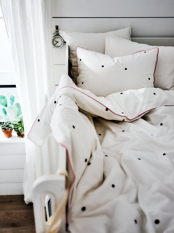 The top of a loft bed by a window with bedding that’s covered in white bed linen with a black spotted detail and a red trim.