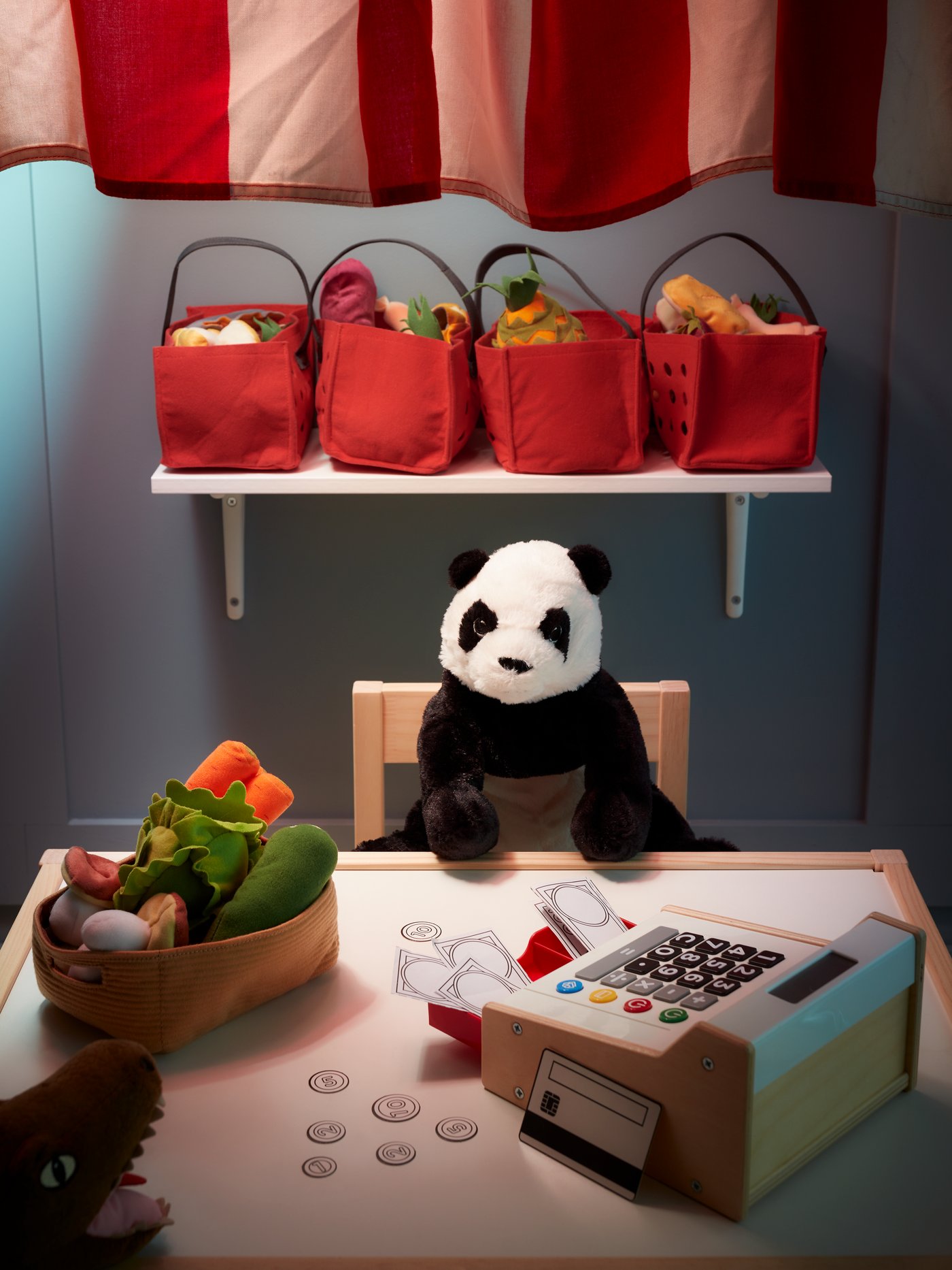 A panda soft toy is placed on a child's chair in front of a table. A toy cash register and a toy basket of vegetables are placed on the table. Above the panda, four toy fruit baskets are placed on a rack.