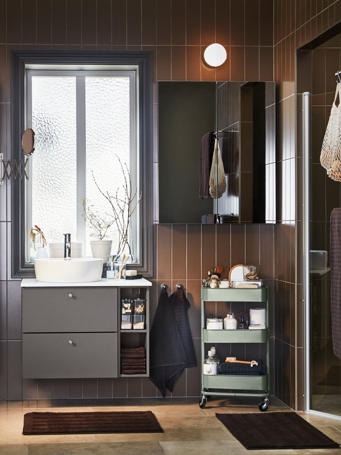 Un baño minimalista convanitorio y mueble mural a juego en tonos grises. Se ven dos afombras de baño en la salida de la ducha y frente al lavamanos.