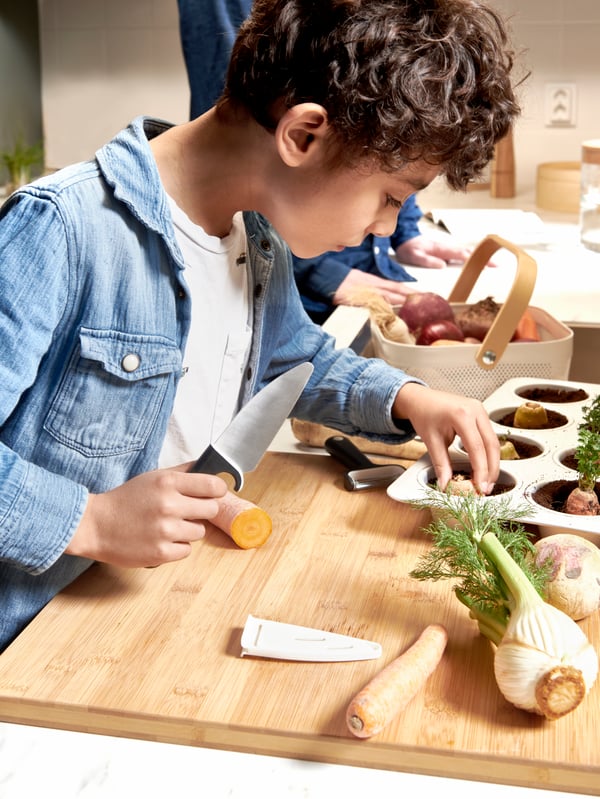 A boy tries to peel real vegetables with the SMÅBIT Knife and peeler on a chopping board.